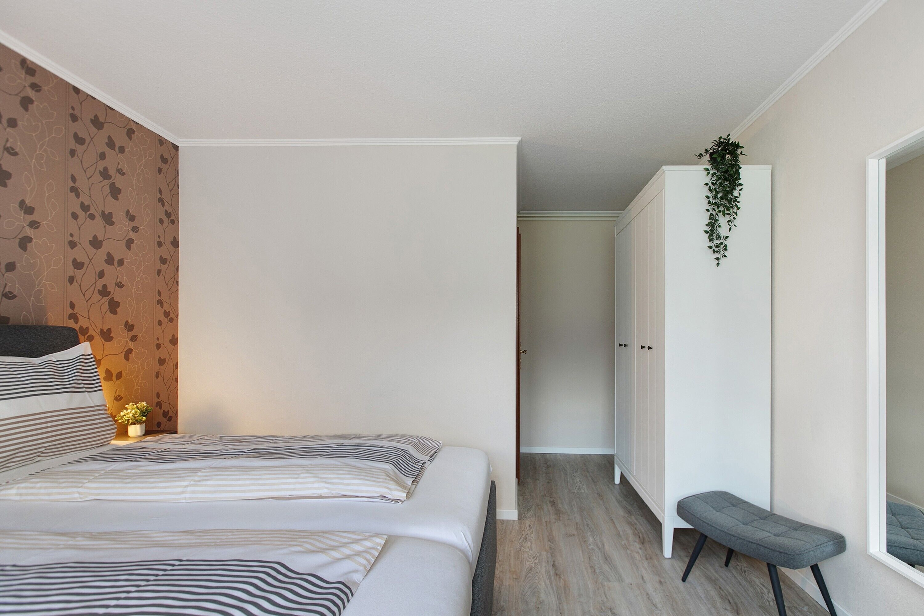 A bedroom with a bed, a bedside table, a bench, a wardrobe, and a hanging plant.