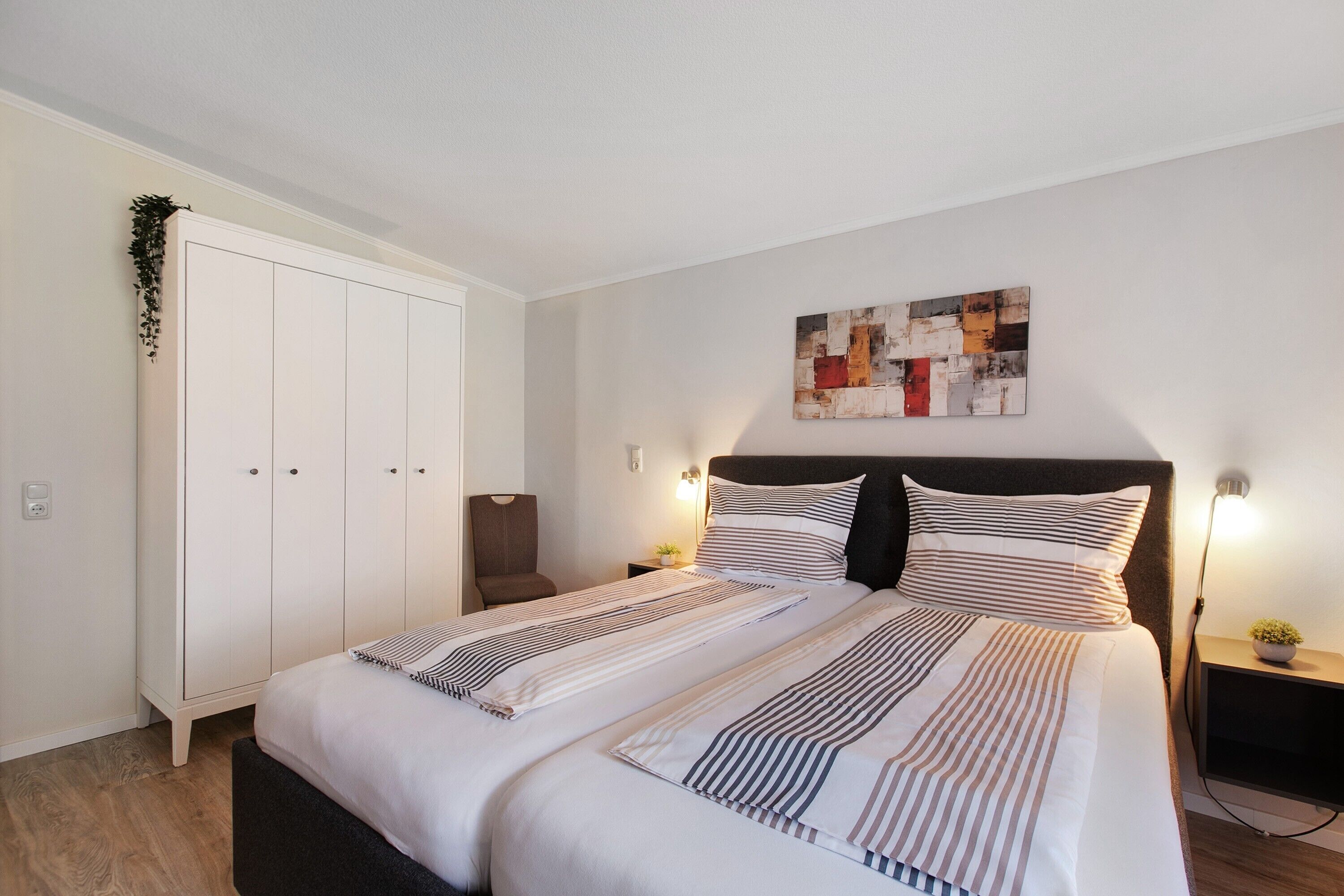 Une chambre à coucher comprenant un lit, une armoire, une chaise, une œuvre d’art murale et une table de chevet avec une lampe.