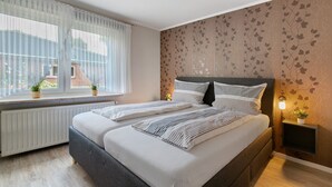 A bedroom with a bed, a window with curtains, a radiator, and a wall with a floral pattern. - Apartment 'Ferienwohnung Sawade' with Private Terrace, Shared Garden and Wi-Fi (Schafstedt)