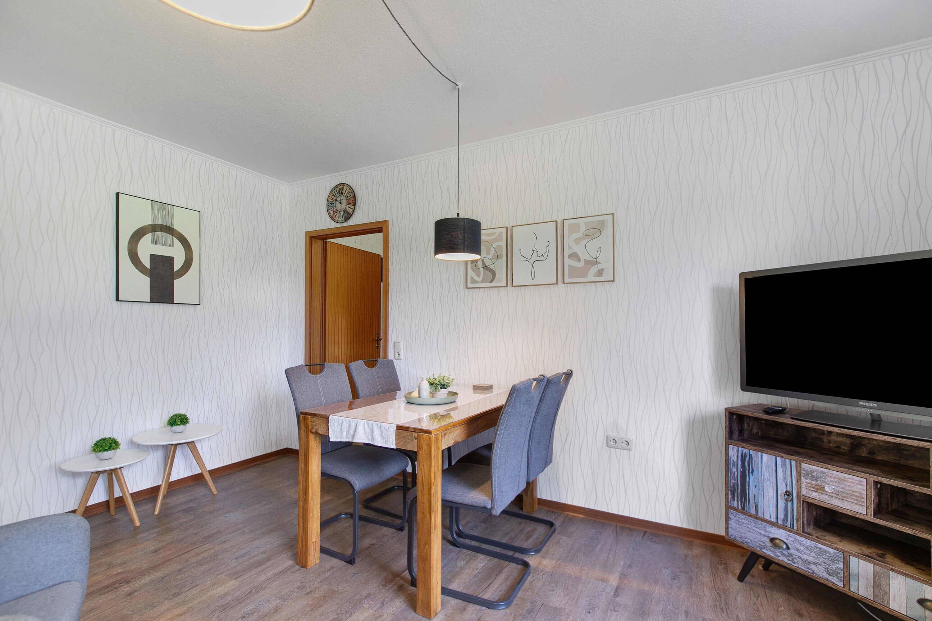 A modern living room with a wooden dining table, grey chairs, a flat-screen TV, and a small side table with a plant.