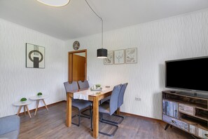 Un salon moderne avec une table à manger en bois, des chaises grises, un téléviseur à écran plat et une petite table d’appoint agrémentée d’une plante.