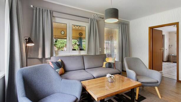 A modern living room with a grey sofa, a wooden coffee table, and a grey armchair. There is a window with curtains, a hanging lamp, and a door leading to another room. - Apartment 'Ferienwohnung Sawade' with Private Terrace, Shared Garden and Wi-Fi (Schafstedt)