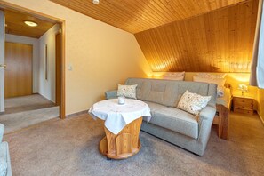 A cozy room with a wooden ceiling, a round wooden table with a white tablecloth, a grey sofa with floral cushions, and a small bedside table with a lamp.