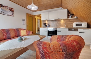 A cozy living area with a sofa, a table with a lace tablecloth, and a patterned armchair. A kitchen with white cabinets and a microwave is visible in the background. - Apartment 'Haus Stoldt, Wohnung 1' with Shared Terrace, Shared Garden and Wi-Fi (Meldorf)