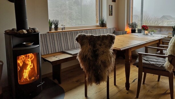 A cozy room with a wood-burning stove, a dining table with chairs, a bench, and a view of trees through a large window. - Apartment 'Am Quellental' with Private Terrace, Private Garden and Wi-Fi (Meinerzhagen)