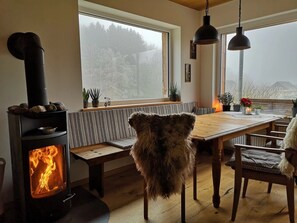 Une pièce chaleureuse avec un poêle à bois, une table à manger avec des chaises, un banc et une vue sur les arbres par une grande fenêtre.