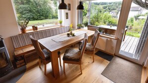 A dining area with a wooden table and chairs, a bench, and a view of greenery through large windows. - Apartment 'Am Quellental' with Private Terrace, Private Garden and Wi-Fi (Meinerzhagen)