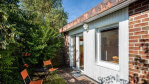 Una casita de ladrillo con puerta blanca y ventana. Cuenta con un pequeño jardín, una silla y una mesa.