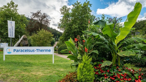 Ein Schild für Paracelsus-Thermo, umgeben von üppigem Grün und blühenden Pflanzen.