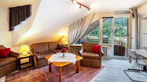 Ein modernes Wohnzimmer mit schrägen Wänden, Blick auf eine grüne Landschaft, einem braunen Ledersofa, einem Holztisch und einem Essbereich mit einem Metallstuhl.