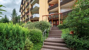 Ein mehrstöckiges Gebäude mit Balkonen, einer Treppe zum Eingang und umgebender Bepflanzung.