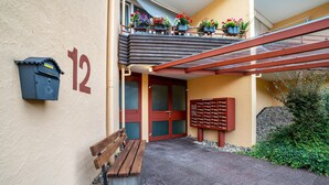 Ein Gebäudeeingang mit Briefkasten, Bank, Tür und einem Balkon mit Blumentöpfen.