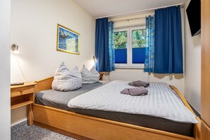 A single bed with a wooden frame, a bedside table, a lamp, a window with blue curtains, and a painting on the wall. - Apartment 'Haus Hügel  - Wohnung 4' with Balcony and Wi-Fi (Binz)