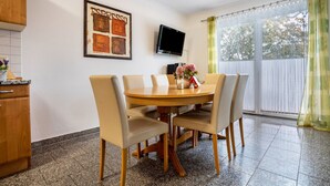 Een eetgedeelte met een ronde tafel, vier stoelen, een aan de muur bevestigde televisie en een raam met gordijnen.