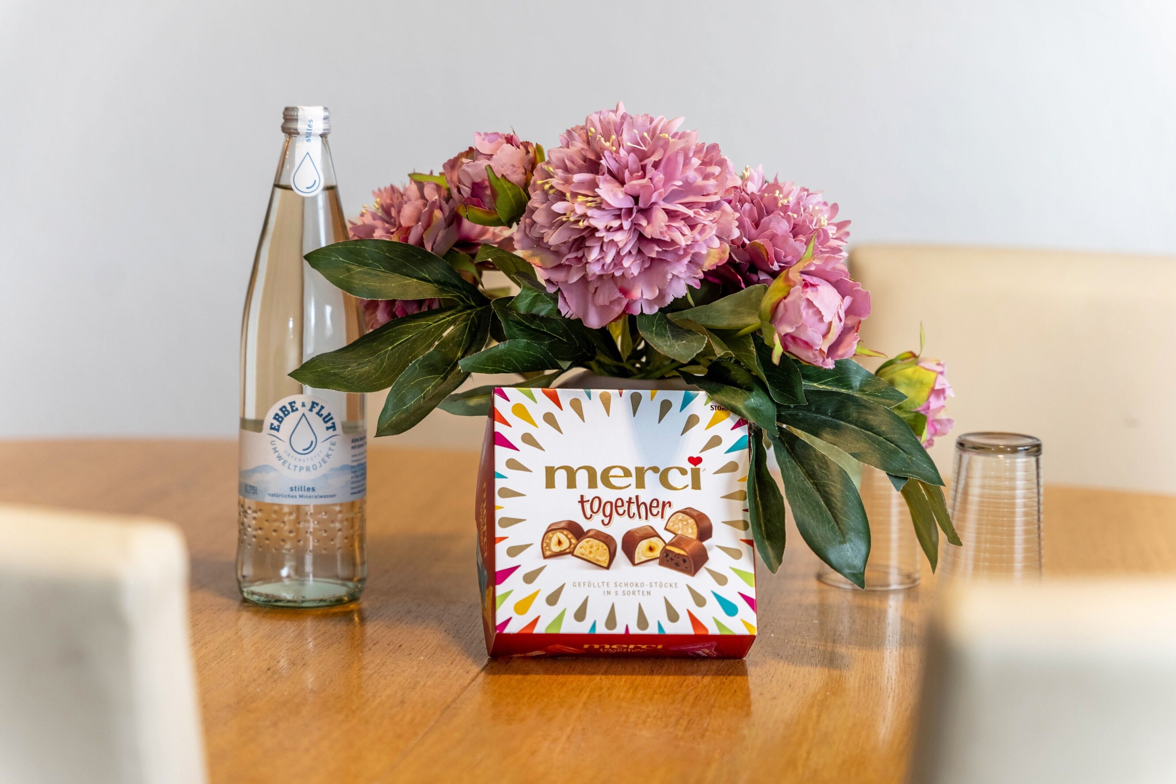 A table with a bottle, a box of chocolates, and a vase of flowers.