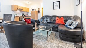 Una sala de estar con un sofá de cuero negro, una mesa de centro de vidrio y una cocina al fondo.