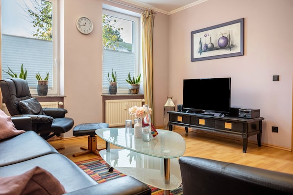 A living room with a leather sofa, a glass coffee table, a TV on a black stand, and potted plants.