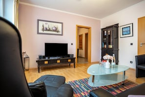 A living room with a flat-screen TV, a glass coffee table, a sofa, a bookshelf, and a door leading to another room.