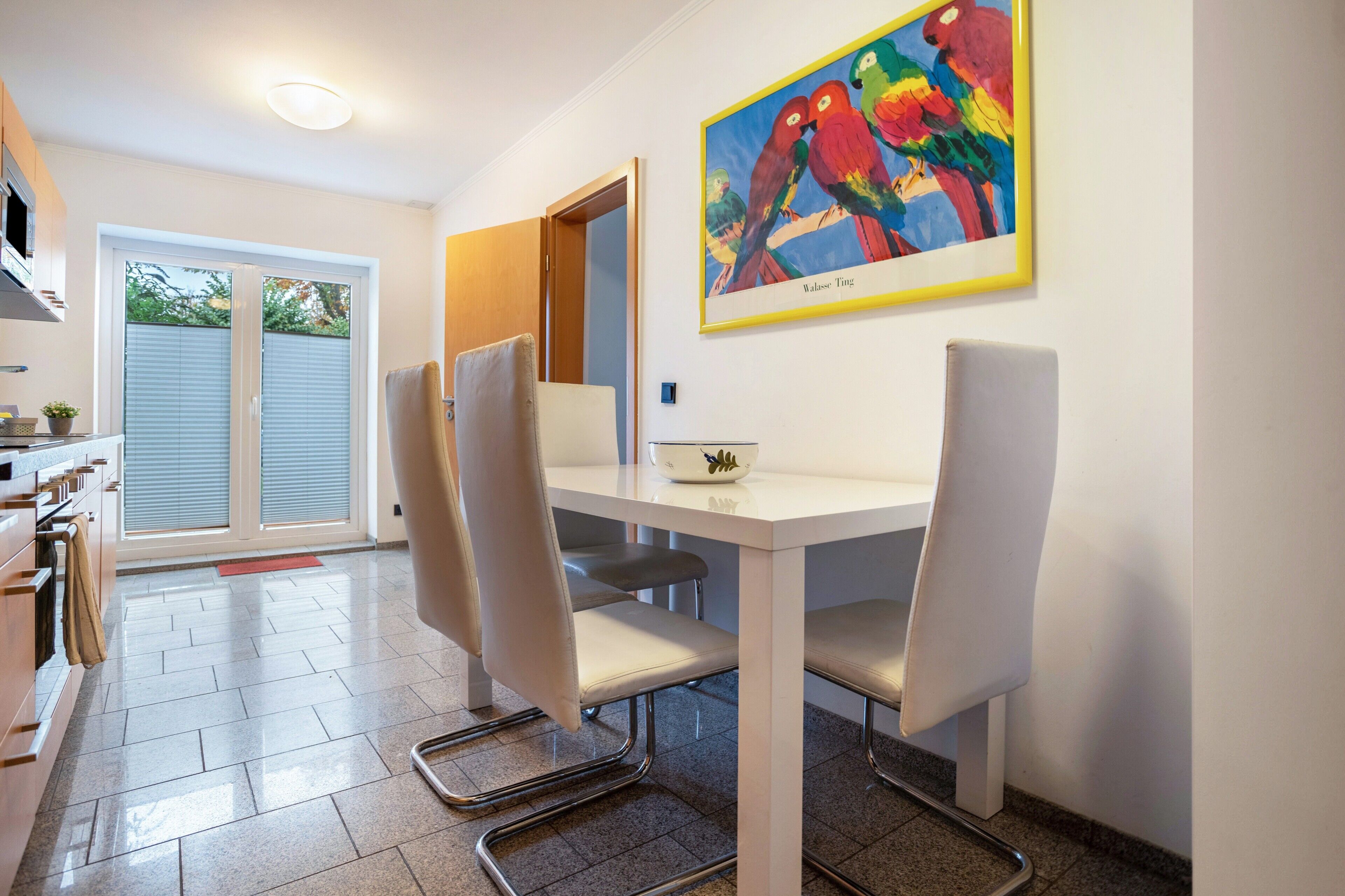 A dining area with a white table, four chairs, a painting of colorful birds, and a view of a sliding door leading to a balcony.