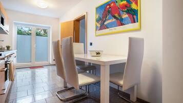 A dining area with a white table, four chairs, a painting of colorful birds, and a view of a sliding door leading to a balcony.