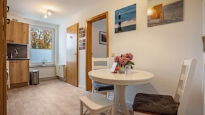 Une pièce comprenant une table ronde blanche, une chaise, une petite poubelle, des meubles en bois, une fenêtre avec des stores et un mur orné de tableaux.