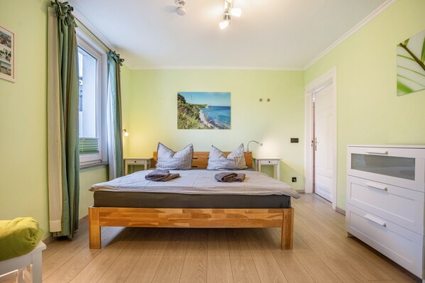 A bedroom with a wooden bed, a nightstand, a bedside table, a window with curtains, a painting on the wall, and a white dresser.