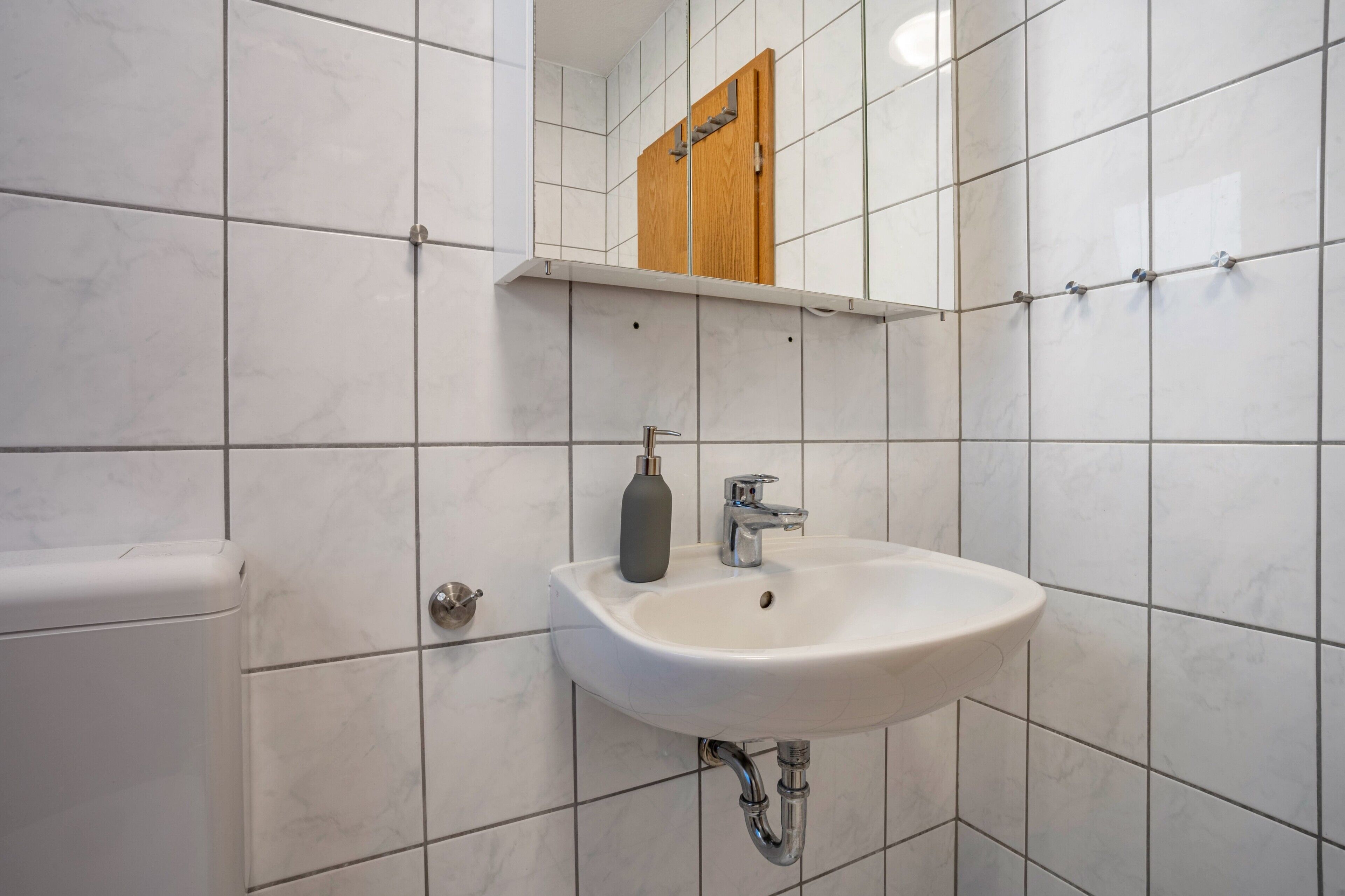 Une salle de bain avec des carreaux blancs, un lavabo blanc, un meuble à pharmacie en bois et des toilettes.
