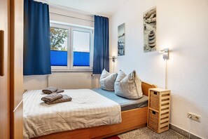 A bedroom with a bed, pillows, a window with curtains, a radiator, a wall-mounted light, and a small wooden storage unit. - Apartment 'Haus Frohsinn - Wohnung 3' with Balcony and Wi-Fi (Binz)