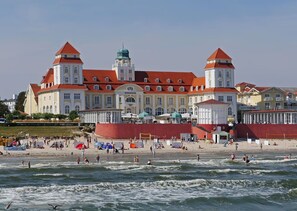 A large building with a red roof and two towers, a beach in front with people and umbrellas, and the ocean in the foreground.