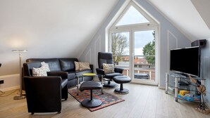 A modern living room with a black leather sofa, a coffee table, a TV on a stand, and a large window. - Apartment 'Bäckerhus - Ferienwohnung 4' with Private Terrace, Shared Garden and Wi-Fi (Wiek)