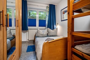 Une chambre avec un lit superposé, un lit simple, un radiateur, une fenêtre avec des rideaux bleus et un tableau encadré au mur.