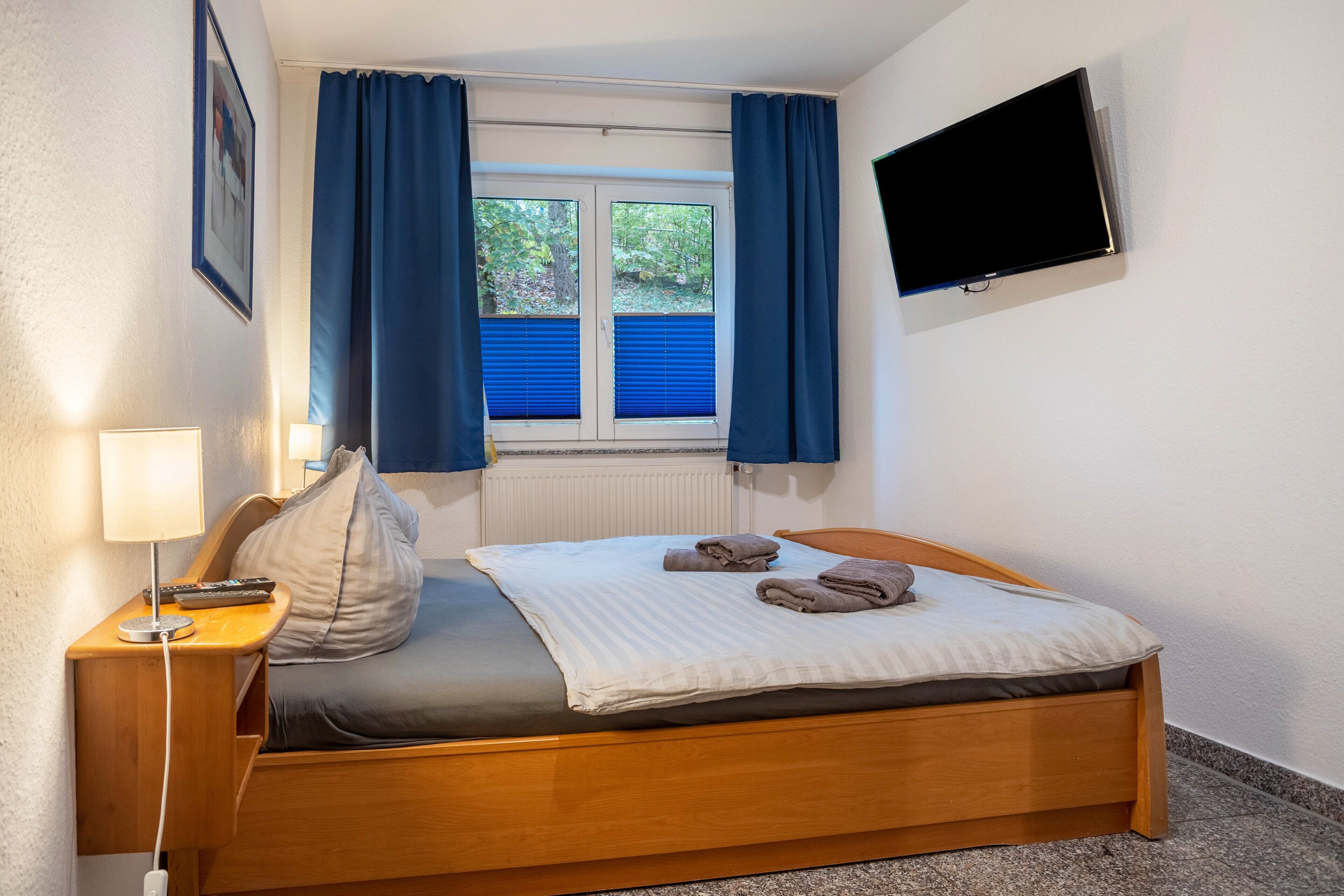 A bedroom with a bed, a TV mounted on the wall, a window with curtains, and a bedside lamp.