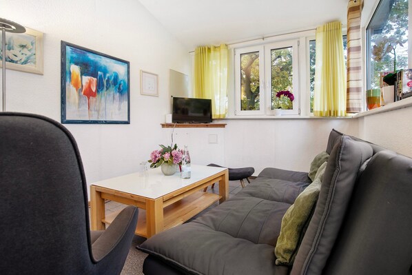 A living room with a sofa, a table with a vase of flowers, a TV on a stand, and a window with curtains.