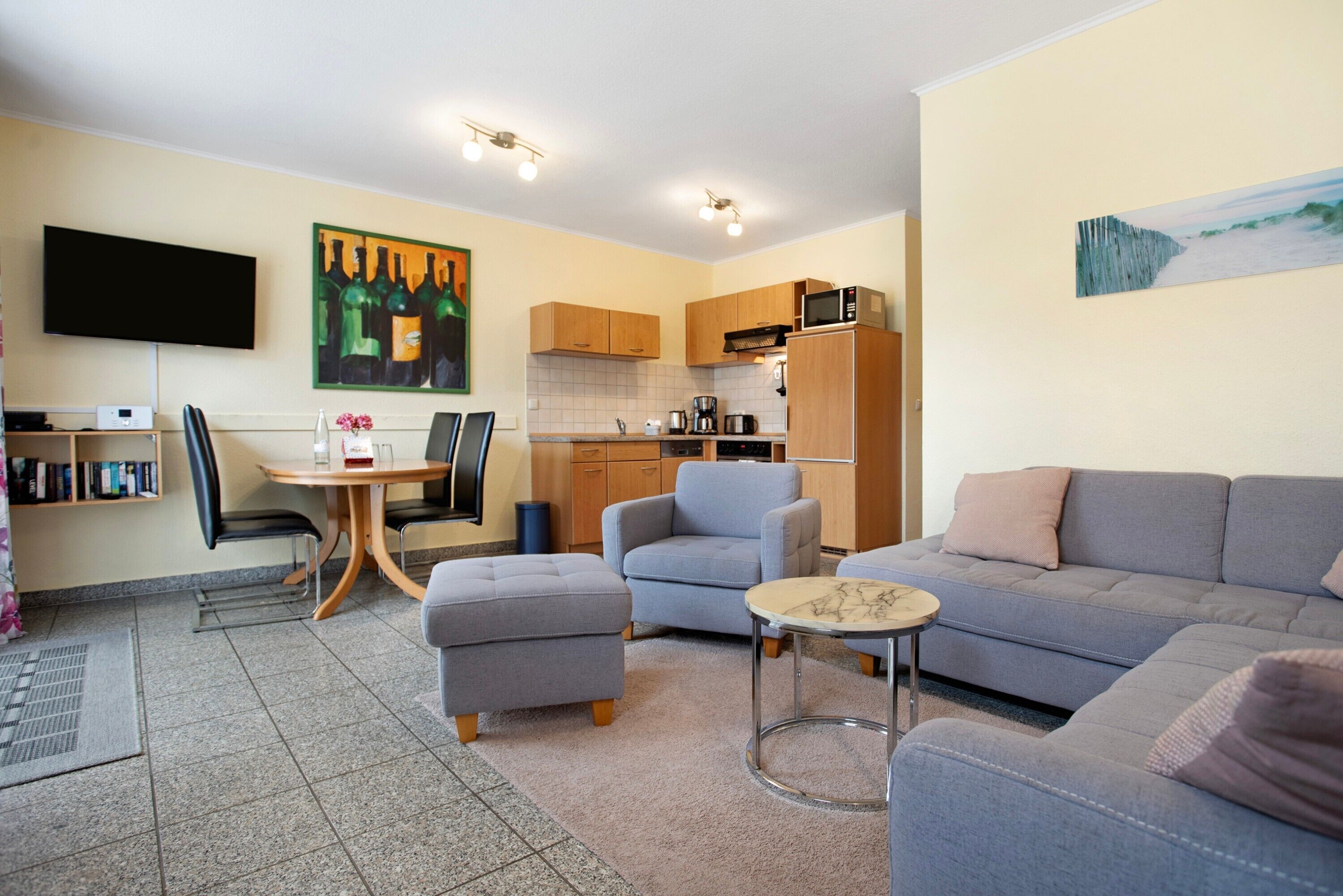 Ein modernes Wohnzimmer mit grauem Sofa, einem kleinen runden Tisch und einem grauen Sessel. Es gibt eine Küchenzeile mit Holzausstattung und einer Mikrowelle. Ein Flachbildfernseher ist über der Sitzecke an der Wand angebracht.