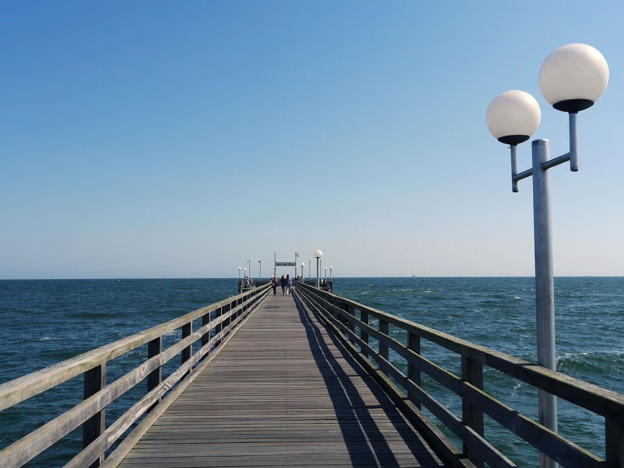 Une jetée en bois s’avançant dans la mer, dotée de rampes et de lampadaires sphériques.