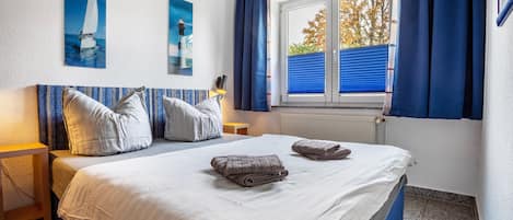 A neatly made bed with pillows and folded towels, a window with blue curtains, and two wall-mounted paintings.