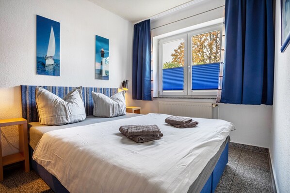 A neatly made bed with pillows and folded towels, a window with blue curtains, and two wall-mounted paintings.