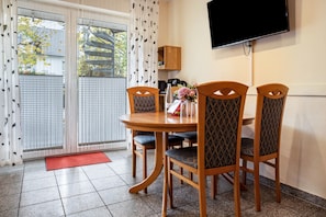 A dining area with a round table, four chairs, a TV mounted on the wall, and a view through a glass door.
