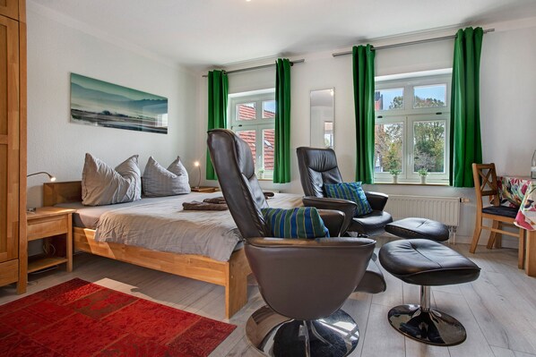 A bedroom with a bed, two leather chairs, a wooden nightstand, a red rug, and a window with green curtains.