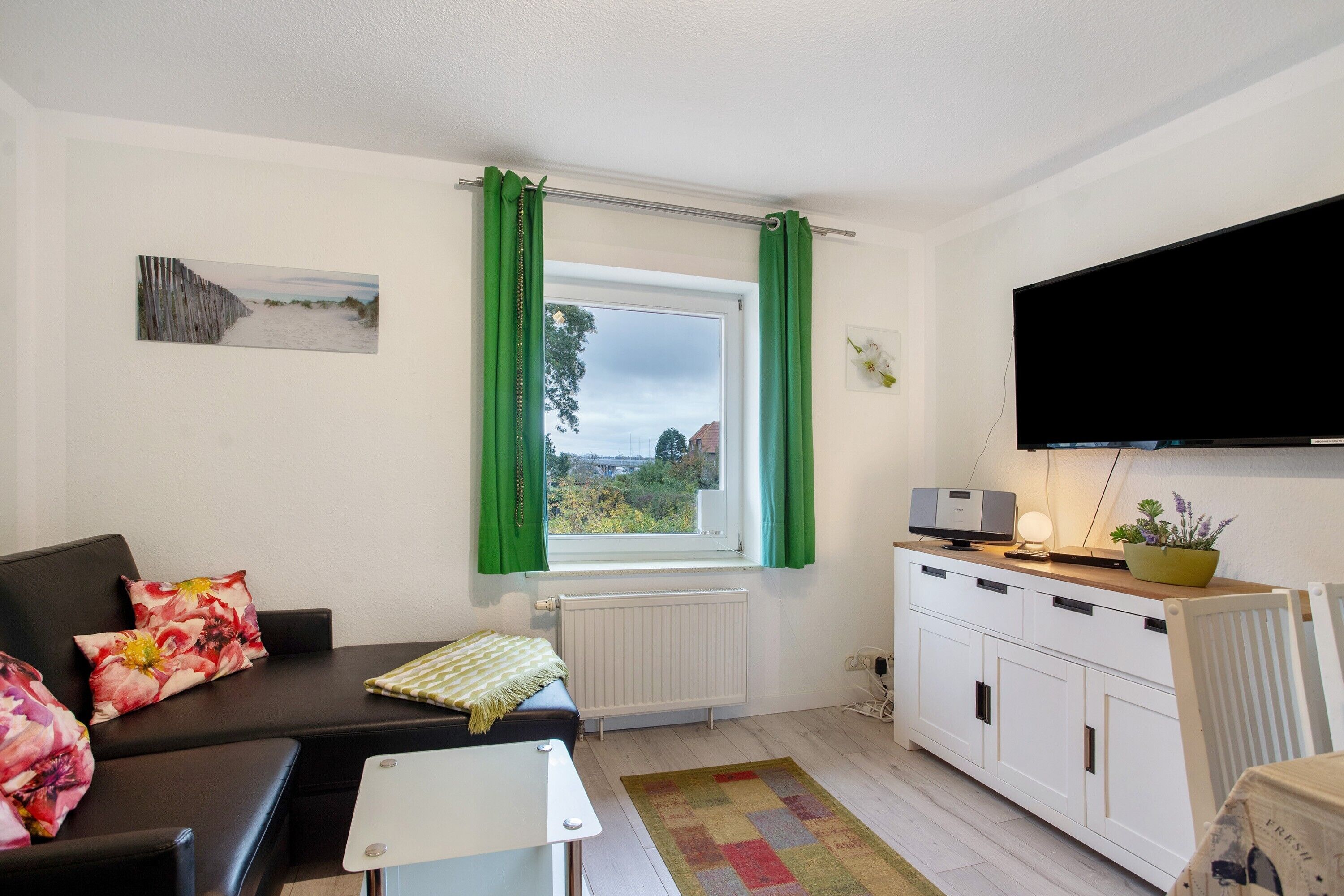 Ein Zimmer mit einer Couch, einem kleinen Tisch, einem Heizkörper, einem Fenster mit Vorhängen, einem Schrank und einem Flachbildfernseher.