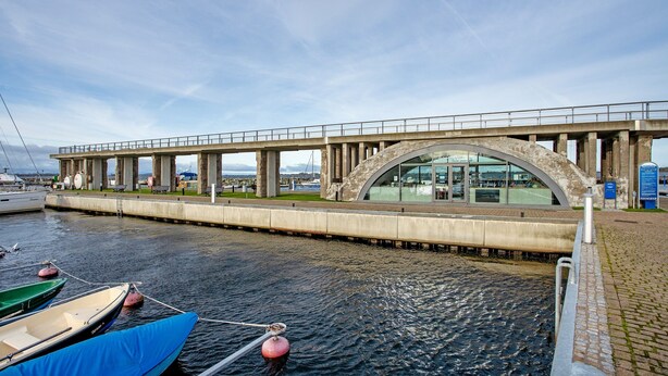 Un puente con una estructura moderna de vidrio, un muelle con barcos amarrados y un paseo marítimo pavimentado.