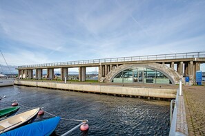 Un puente con una estructura moderna de vidrio, un muelle con barcos amarrados y un paseo marítimo pavimentado.