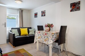Un salon avec un canapé noir, une table à manger recouverte d’une nappe à motifs floraux et une fenêtre avec des rideaux.