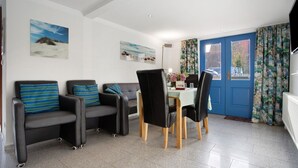 Een kamer met een eettafel, twee banken met gestreepte kussens, een blauwe deur en een schilderij van een strand aan de muur.