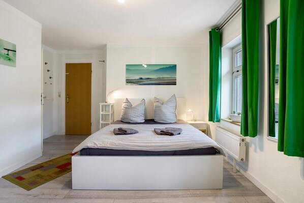 A bedroom with a bed, pillows, a painting, a window with curtains, a radiator, and a door.