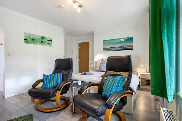 Quarto com duas poltronas de couro e estrutura de madeira, cama com colchão branco e almofadas listradas, uma mesinha com uma xícara, e um quadro na parede.