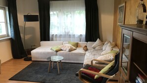 Una sala de estar con un sofá, una mesita redonda, una silla, una estantería y una ventana con cortinas.