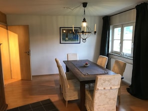 A dining area with a wooden table, four chairs, a chandelier, a framed picture on the wall, and a window with curtains.