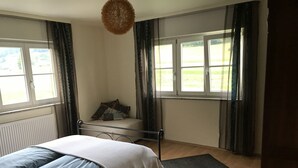 Un dormitorio con cama, mesita de noche, radiador, ventana con cortinas y vistas al exterior.