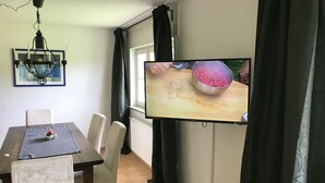 Un comedor con una mesa, sillas, un televisor montado en la pared y un ventanal con cortinas.